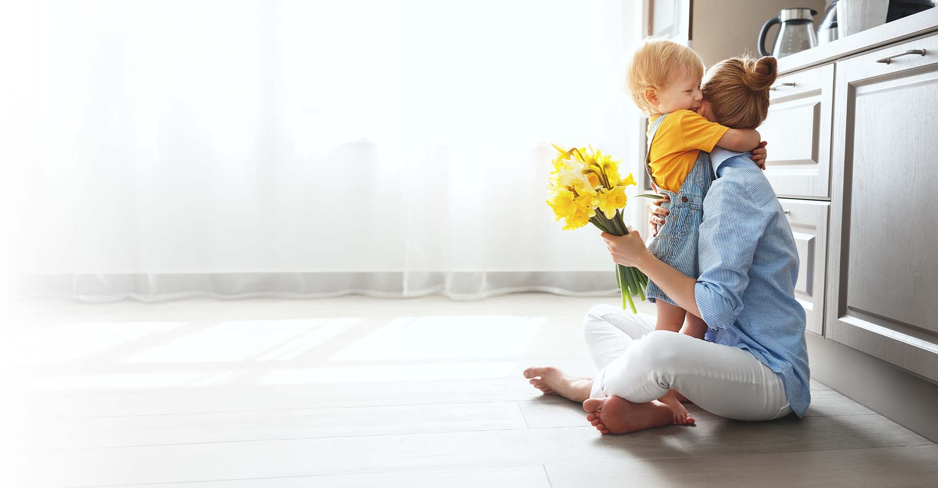 Mother hugging son sitting on kitchen floor holding yellow flowers