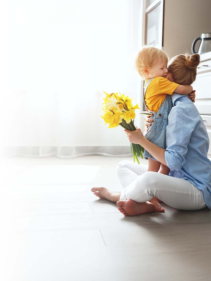Mother hugging son sitting on kitchen floor holding yellow flowers